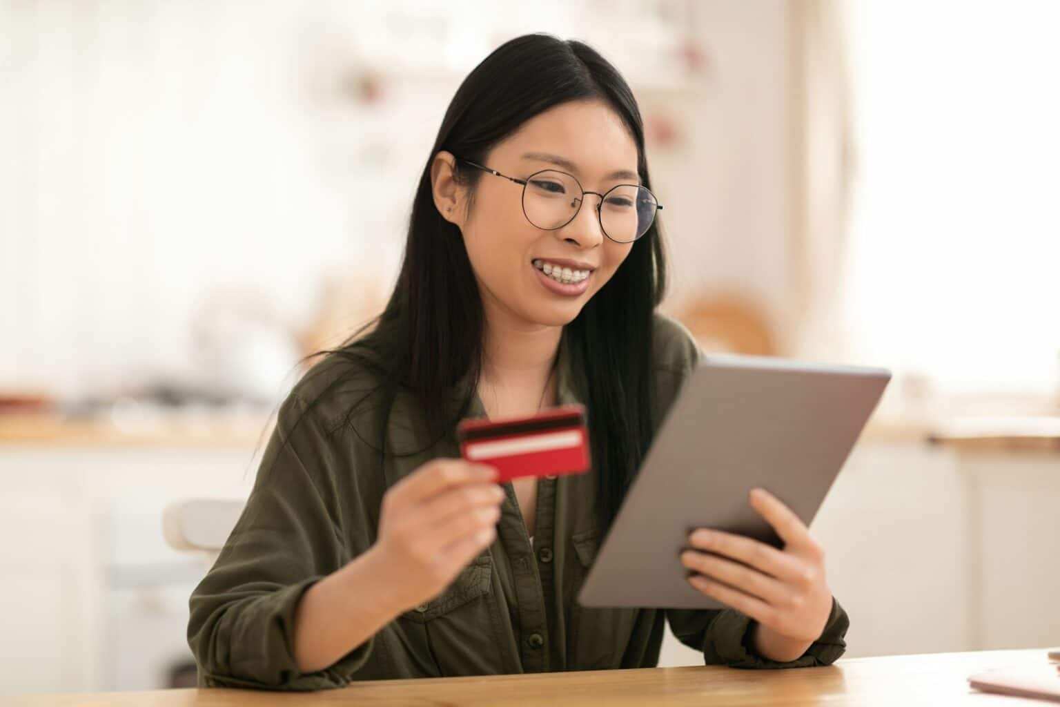 Avis Hello bank! : nos conseils avant d’ouvrir un compte Smiling asian woman using tablet and bank card at kitchen