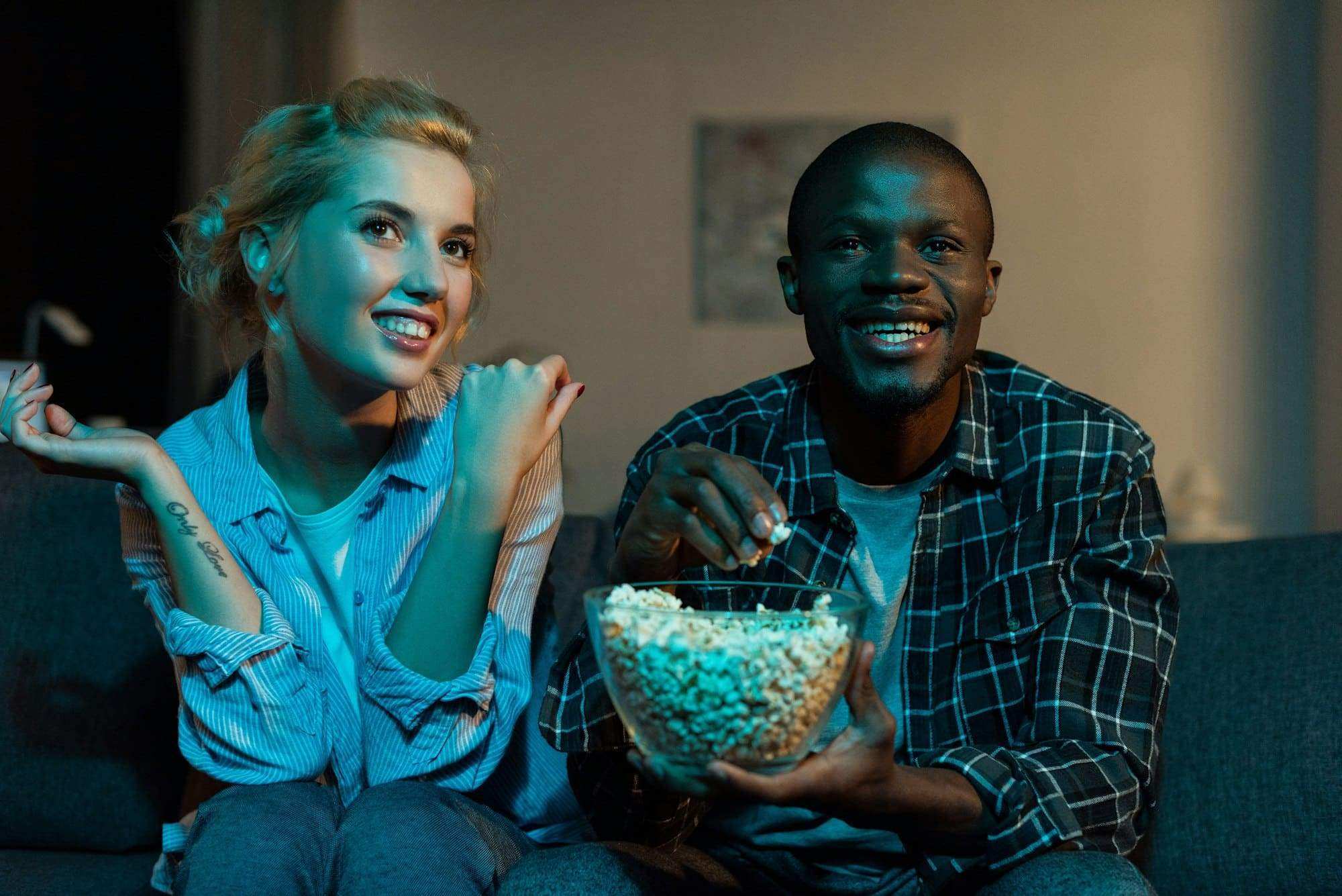 portrait of cheerful multiethnic couple watching tv together on sofa at home