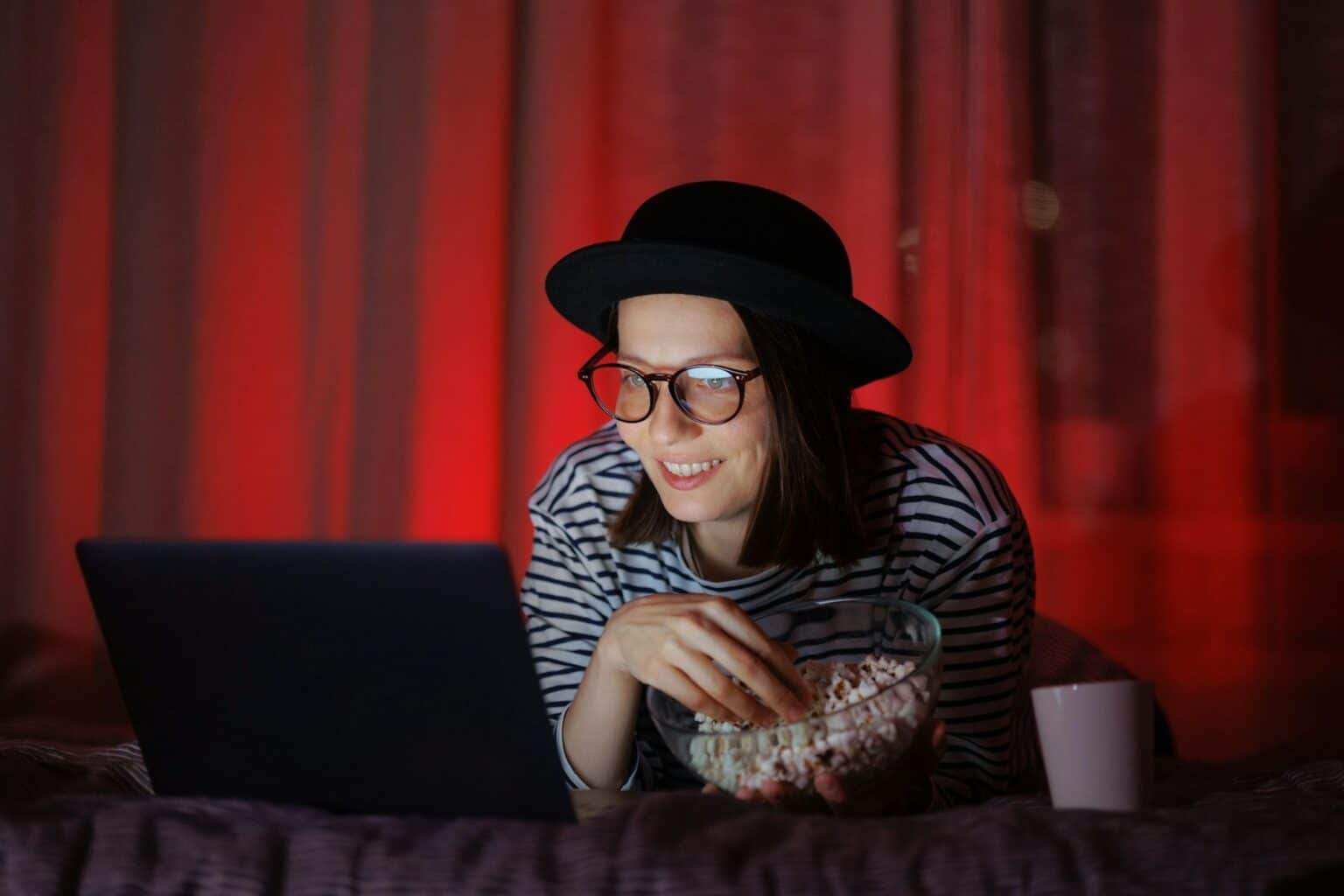 woman in a dark room with a hat at home watching TV or a movie on a laptop and eating popcorn.
