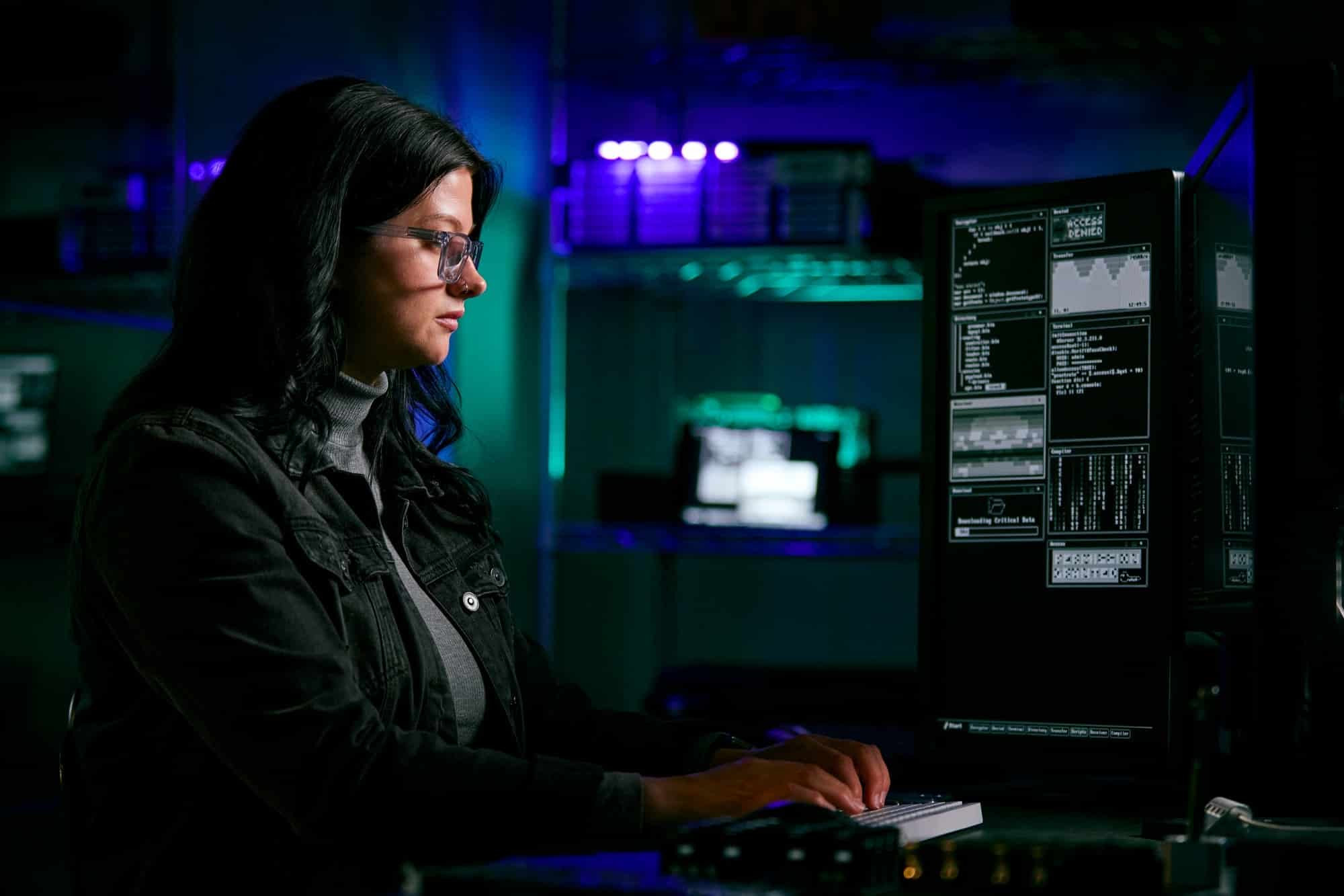 Low Key Lighting Shot Of Female Computer Hacker Sitting In Front Of Screens Breaching Cyber Security