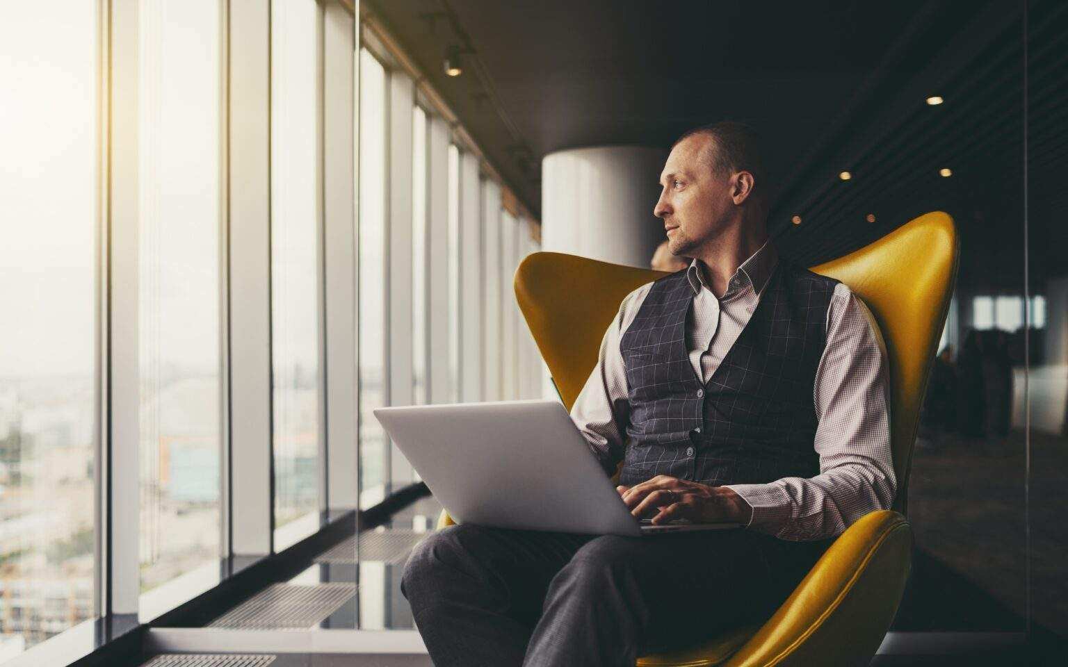 A businessman on an armchair with a laptop, looking out the window