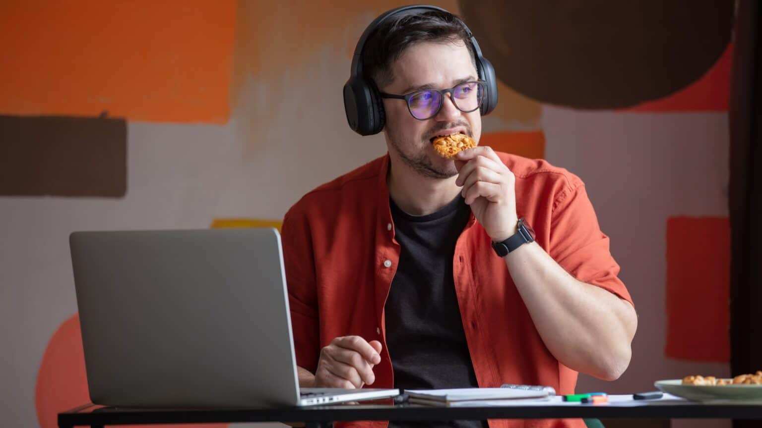 Man in headphones working in home office and eating a cookie at table