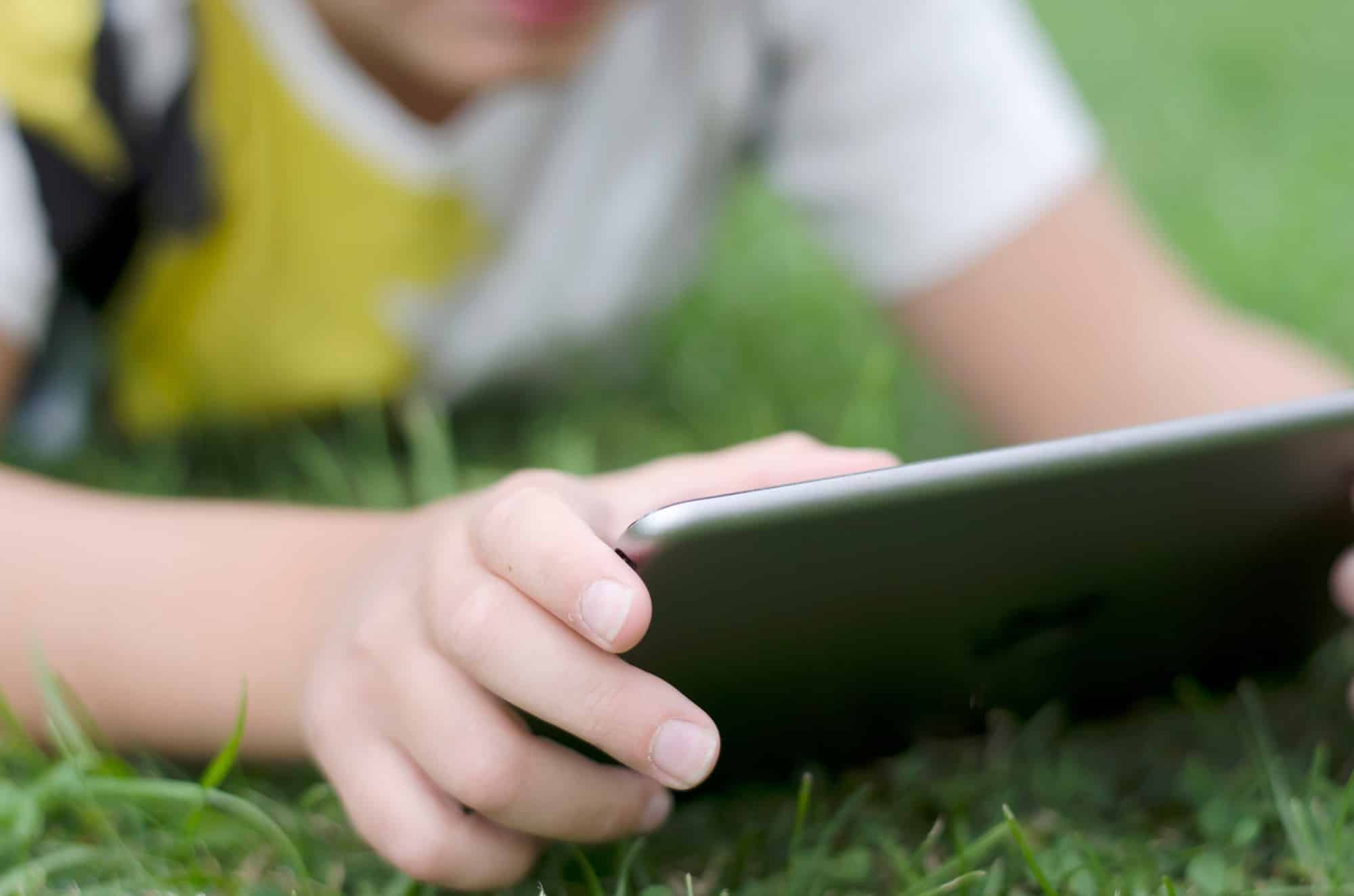 A kid using iPad lying on the green grass backyard relaxing gaming having fun happiness vacations