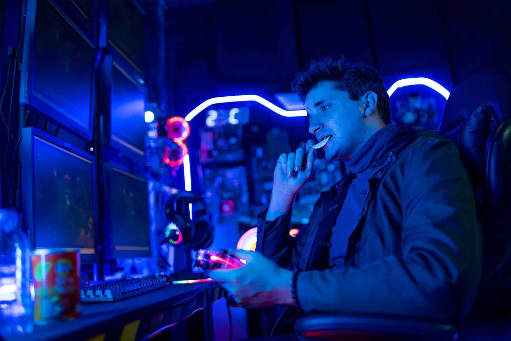 Caucasian gamer man eat potatoes chip during play game in the room with multi-color light