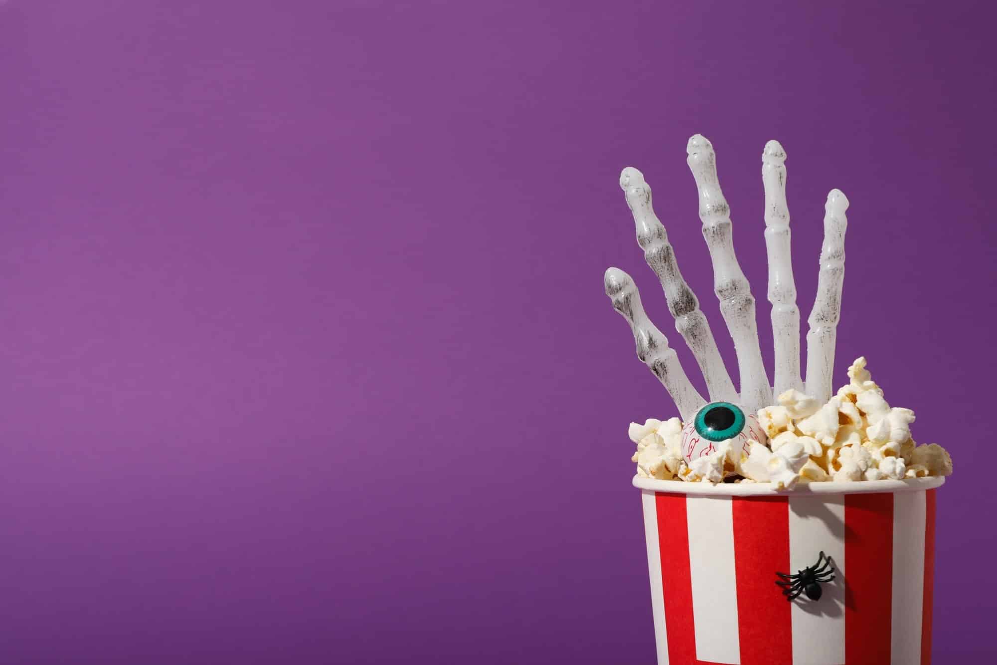 Paper cup with popcorn and Halloween decorations on purple background, space for text