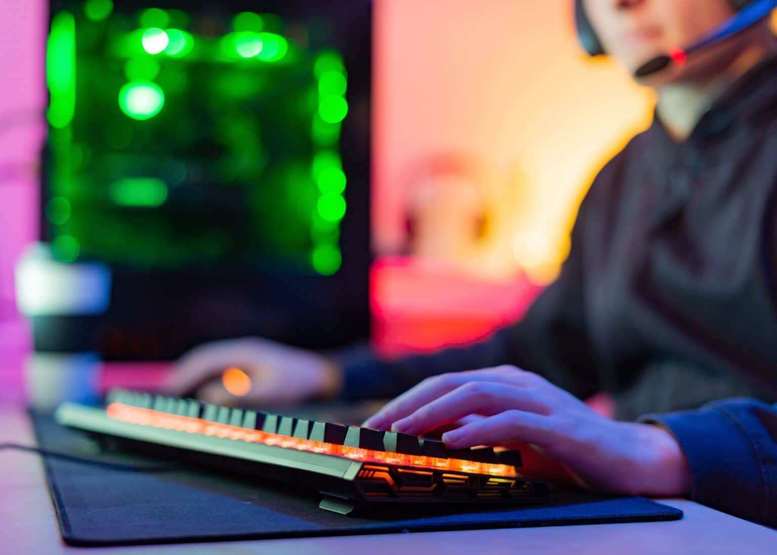 Unrecognizable young gamer with hands on illuminated keyboard in room with colored background