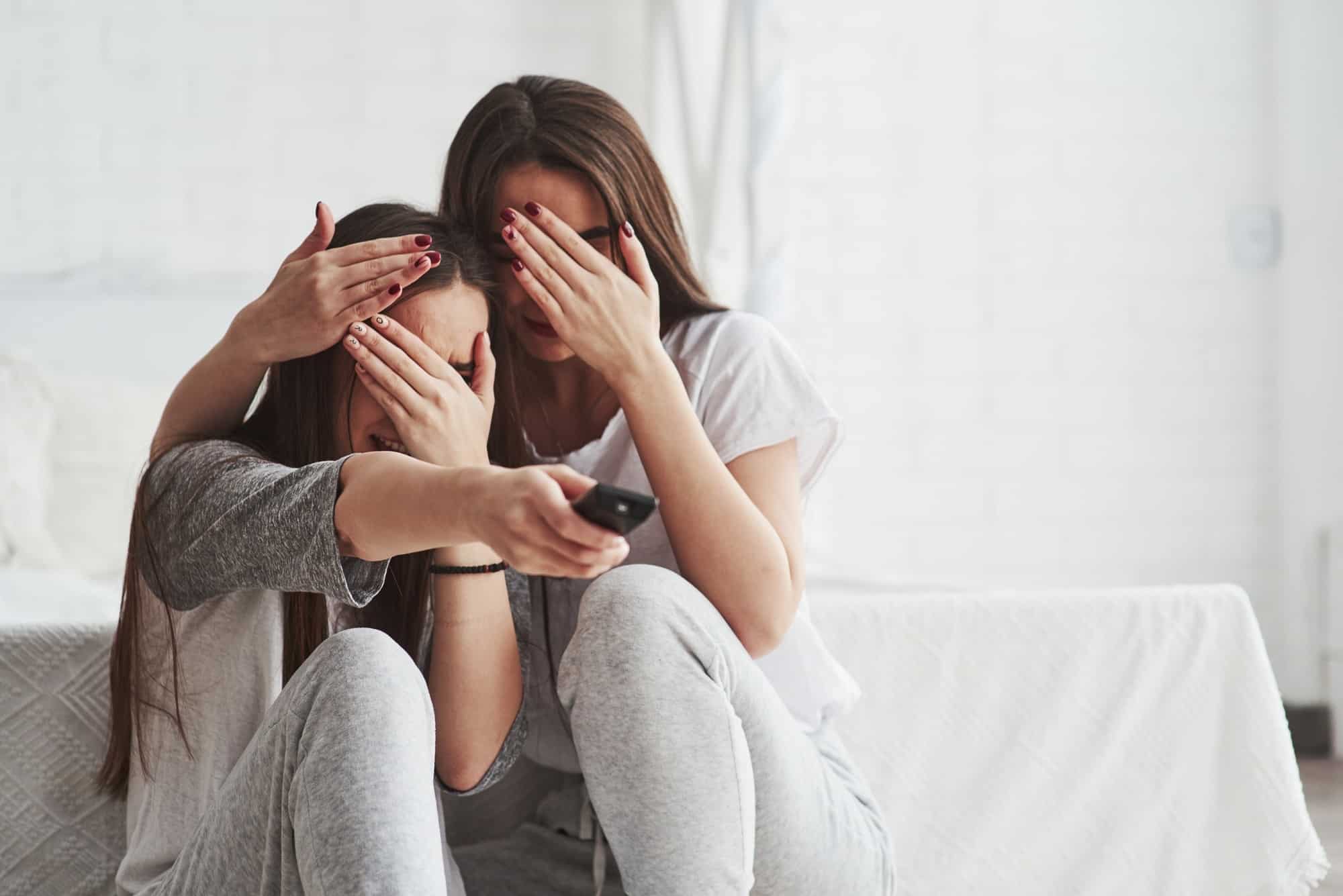 Sisters have fun watching TV while sits on the floor of beautiful bedroom at daytime