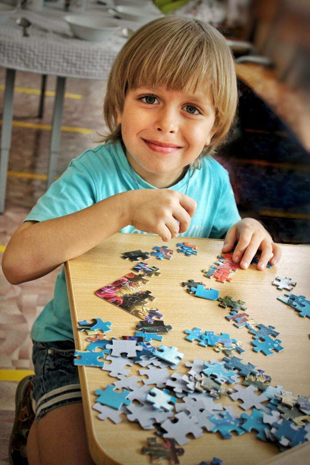 A cute smiling blond boy of 6 years old is sitting at the table and collecting a picture from puzzle