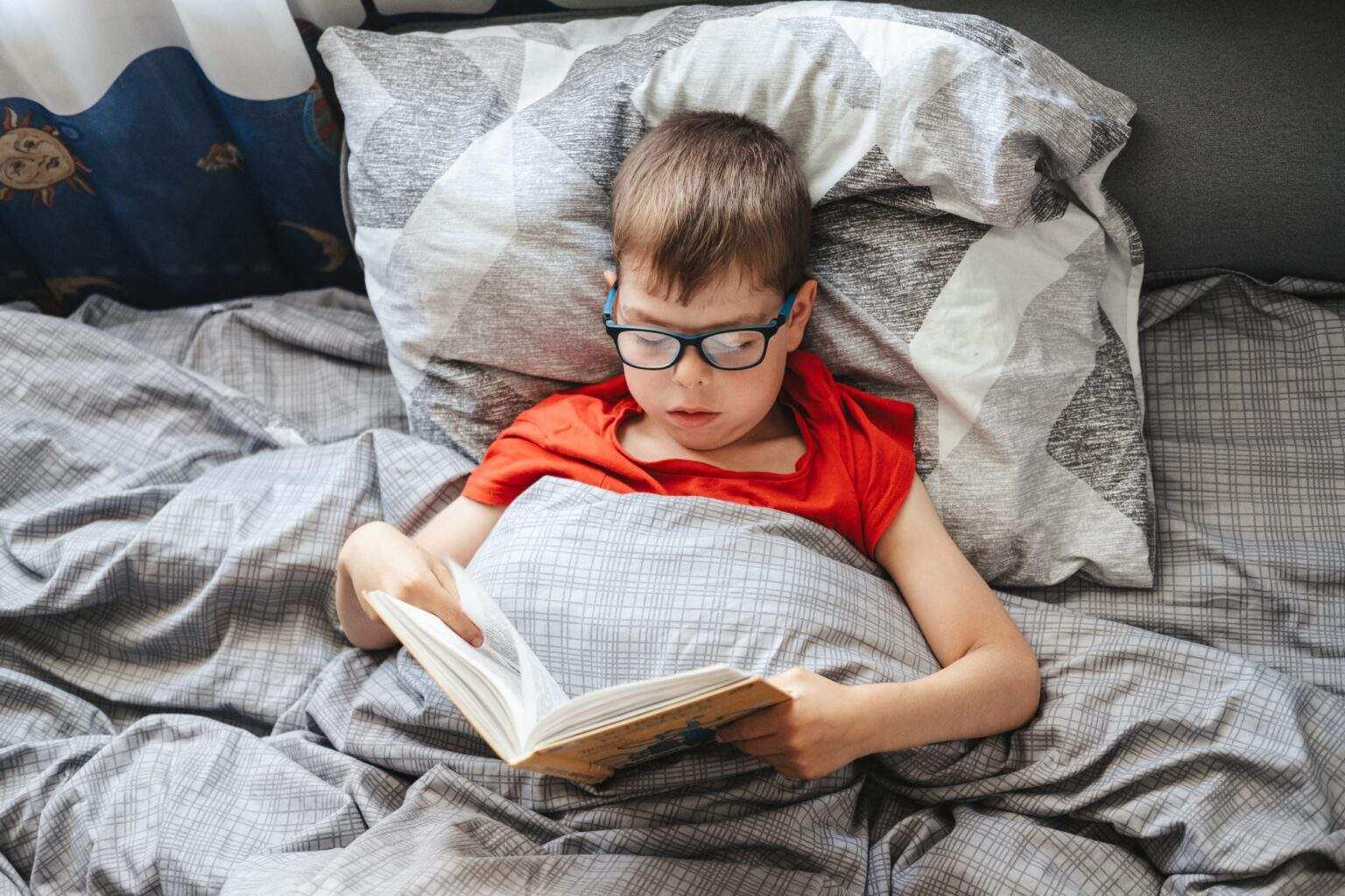 boy with glasses lies on a bed under a blanket and reads a book .child is reading a book in bed.